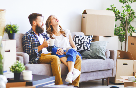 Young couple sitting on couch in their new home, smiling and surrounded by moving boxes