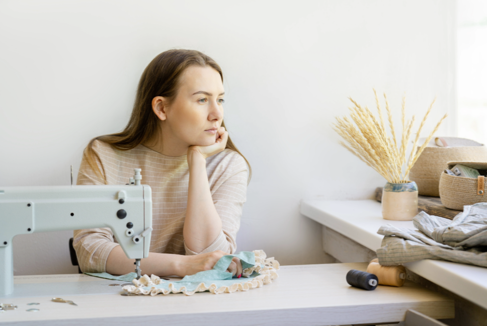 Woman sitting at a sewing machine, reflecting while working on a handmade garment—representing side hustle income like Etsy or craft-based business.